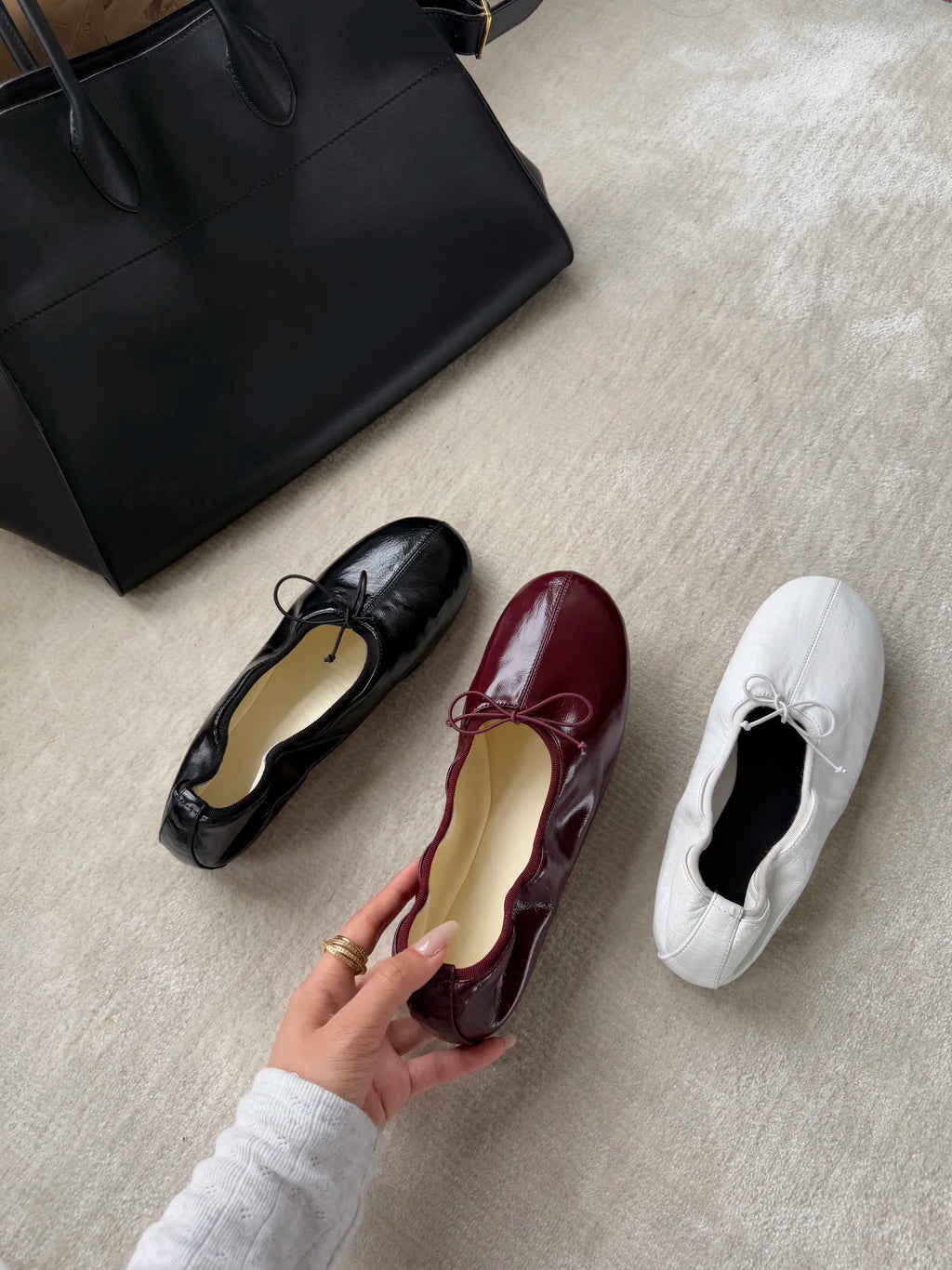 Three shoes (black, red, white) on a light gray surface with a black handbag.