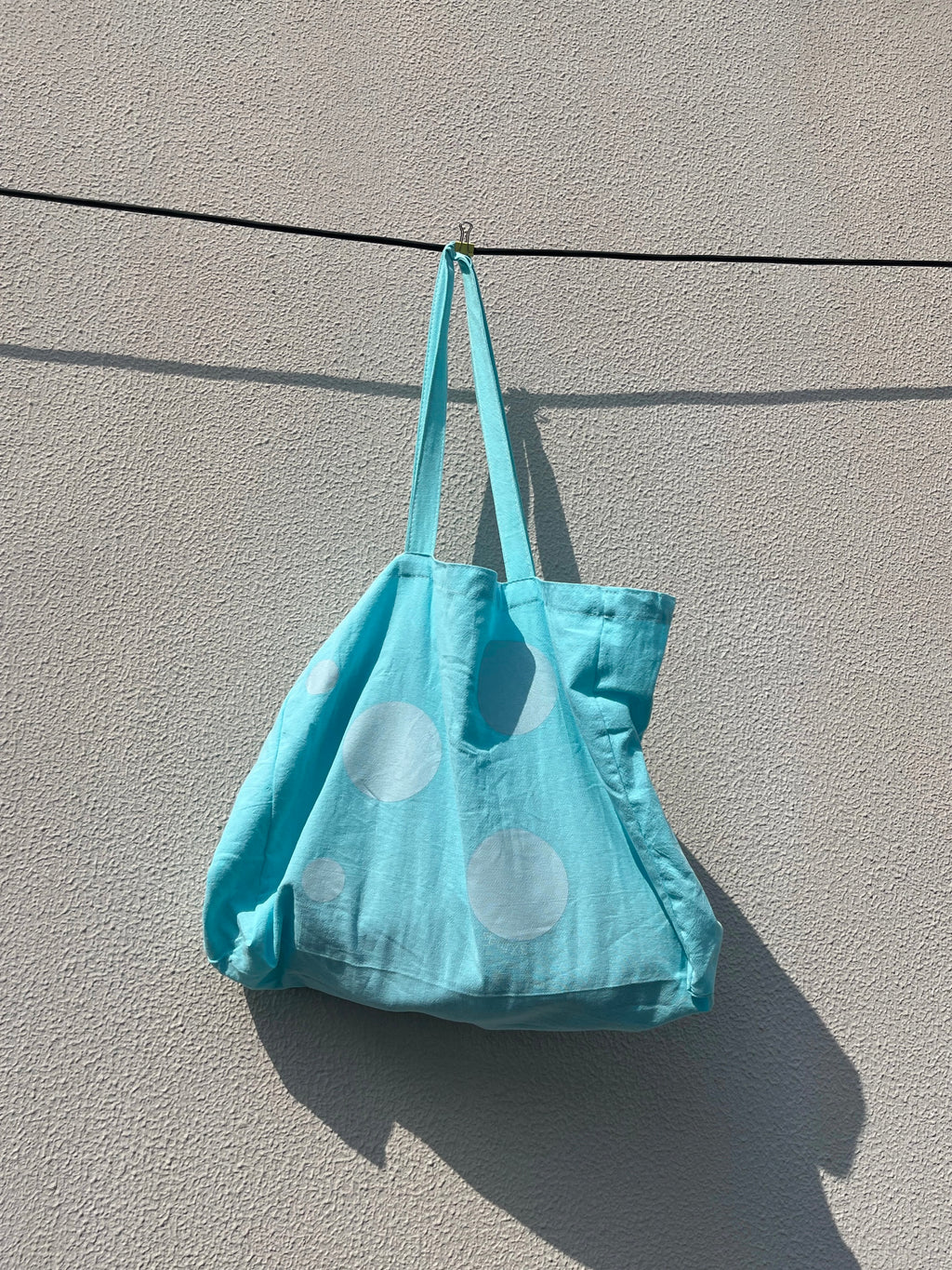 Blue tote bag with white polka dots hanging on a wire against a gray wall.