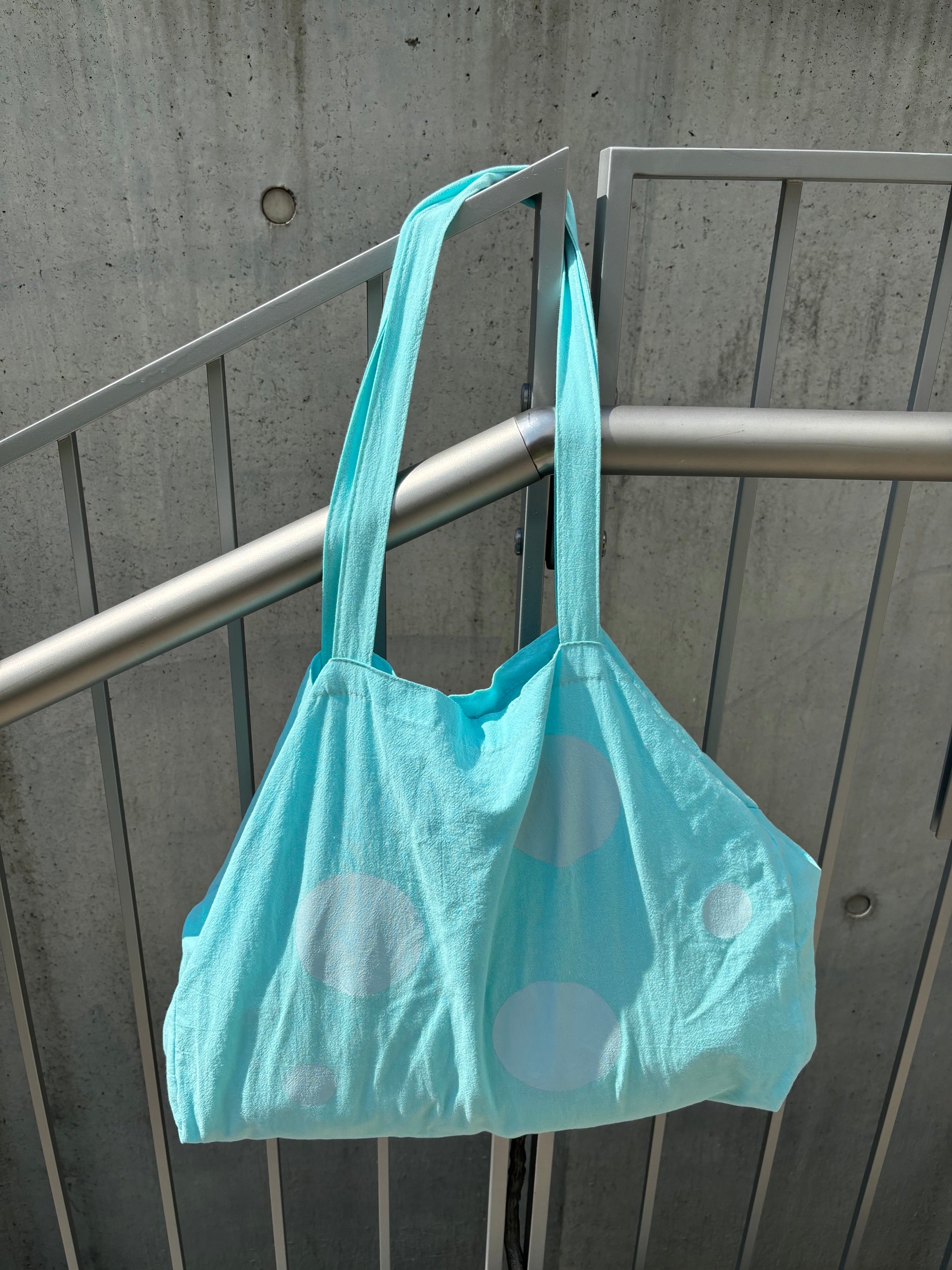 Light blue tote bag with white polka dots hanging on a metal railing.